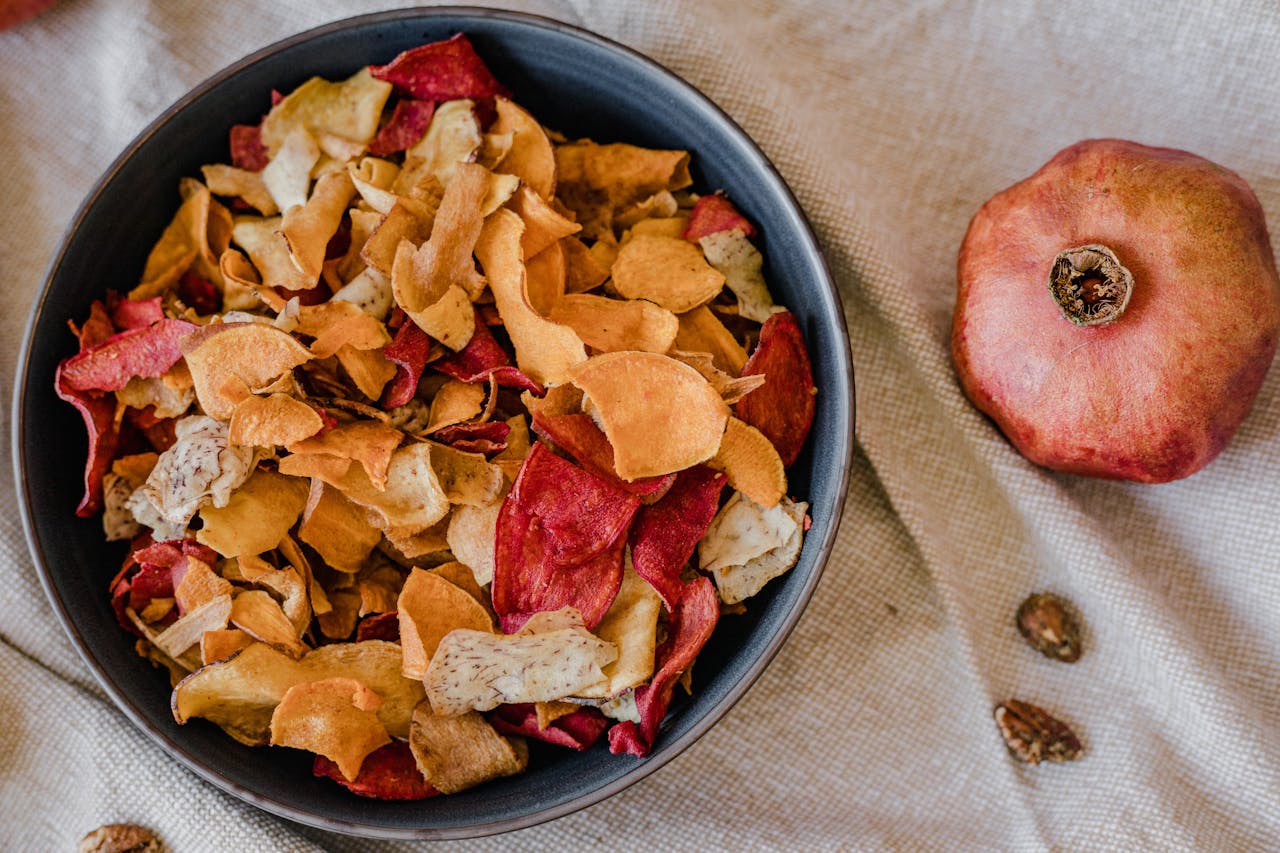 A stylish presentation of healthy dried fruit chips in a bowl with a pomegranate on textile background.